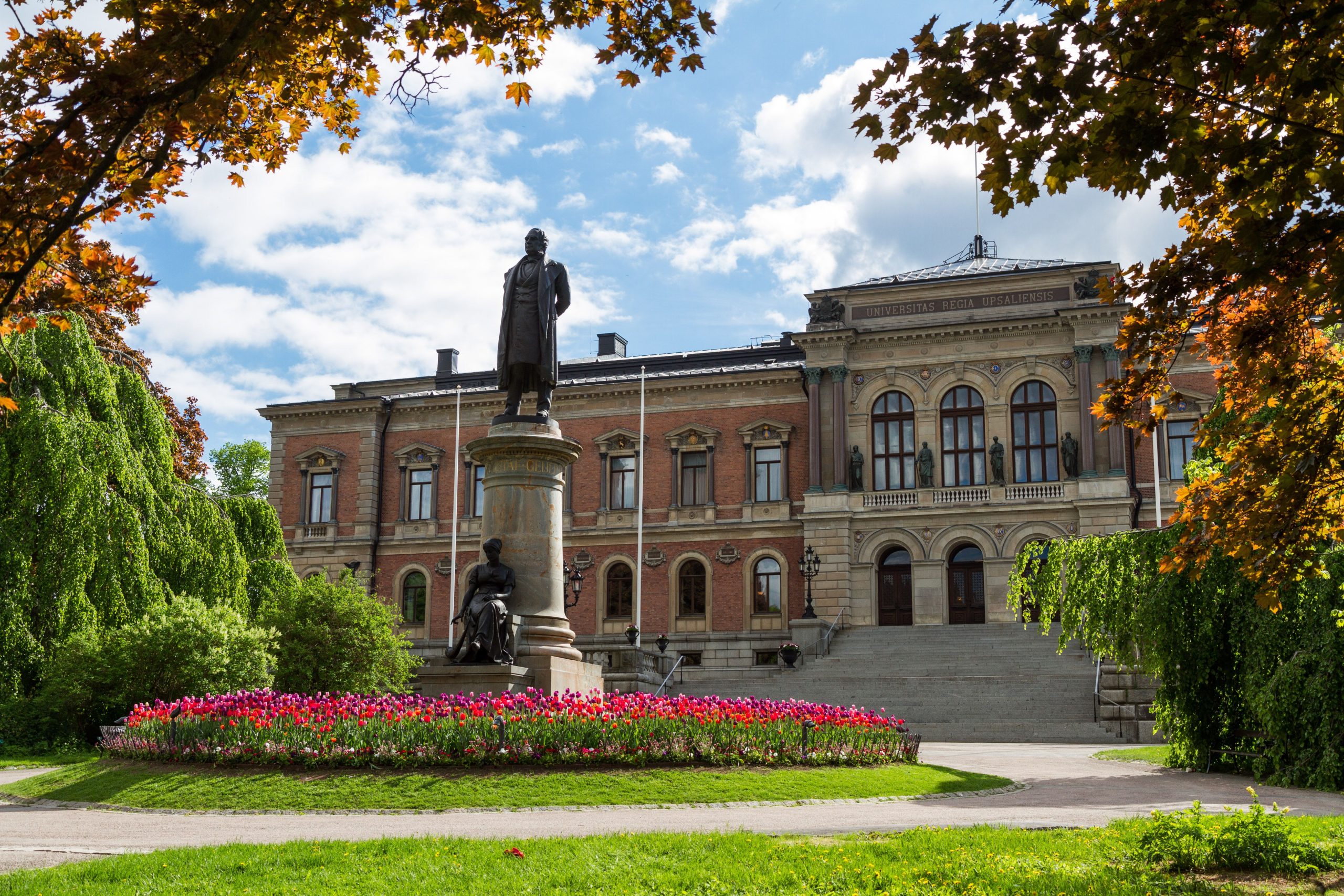 Uppsala University Main building, "Universitetshuset". A statue, flowers and trees, in front of a 19th century building. Photo.