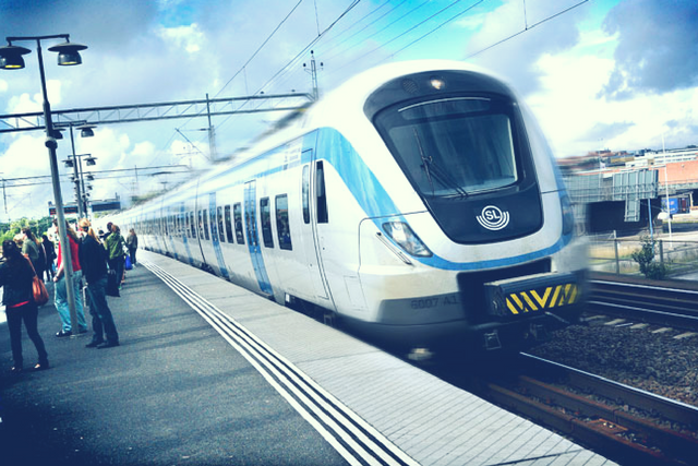 Front of a train passing a platform with people waiting. Photo.