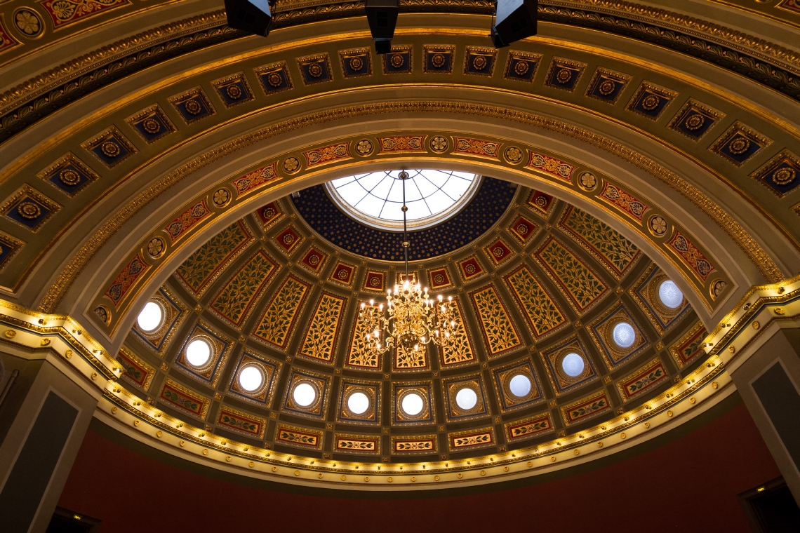 A chandelier hanging from a ceiling window, surrounded by a patterned ceiling vault. Photo.